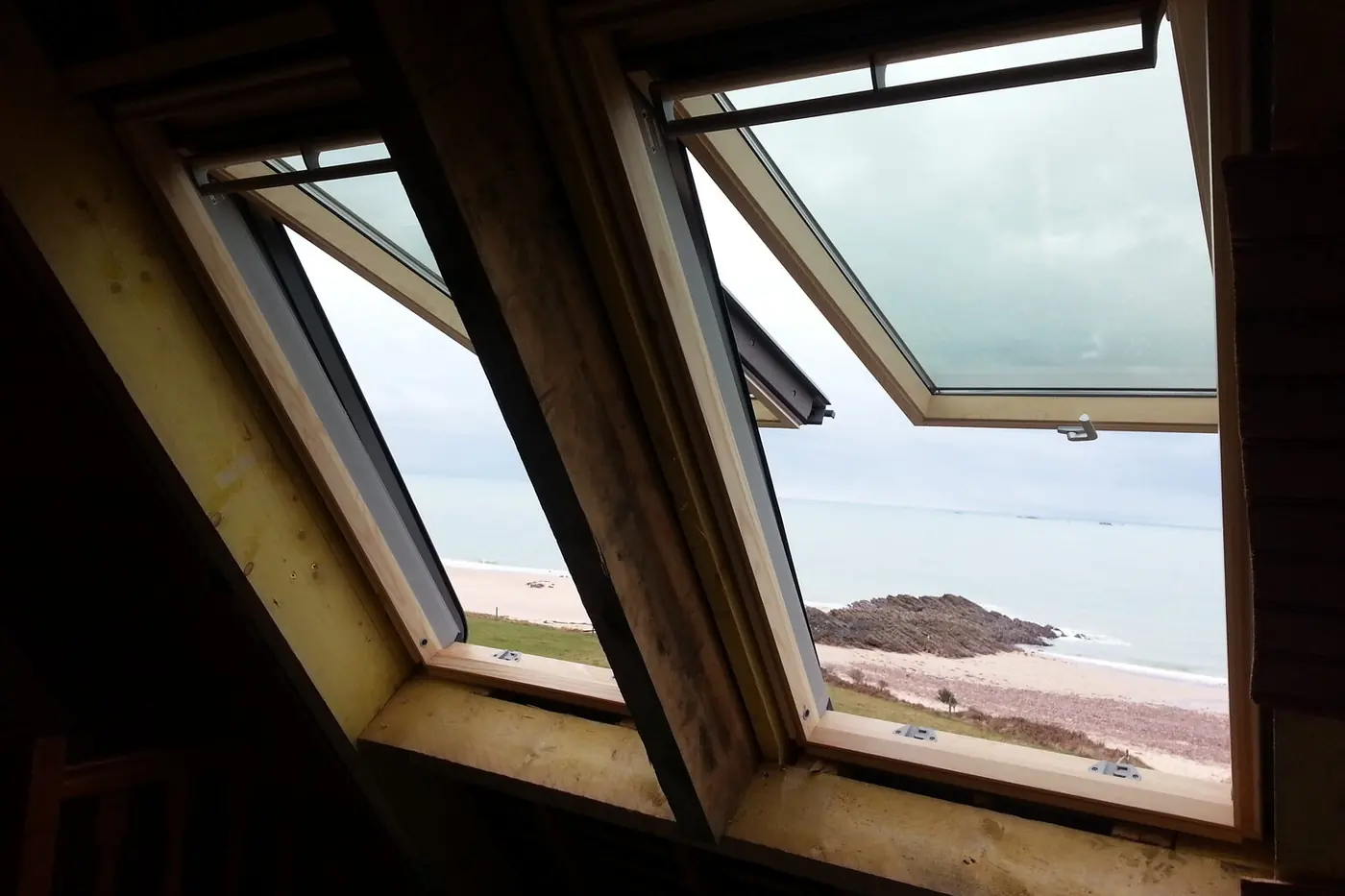 Deux fenêtres de toit ouvertes dans un rampant, vues de l'intérieur d'un comble : au-dehors, la plage, les rochers et la mer
