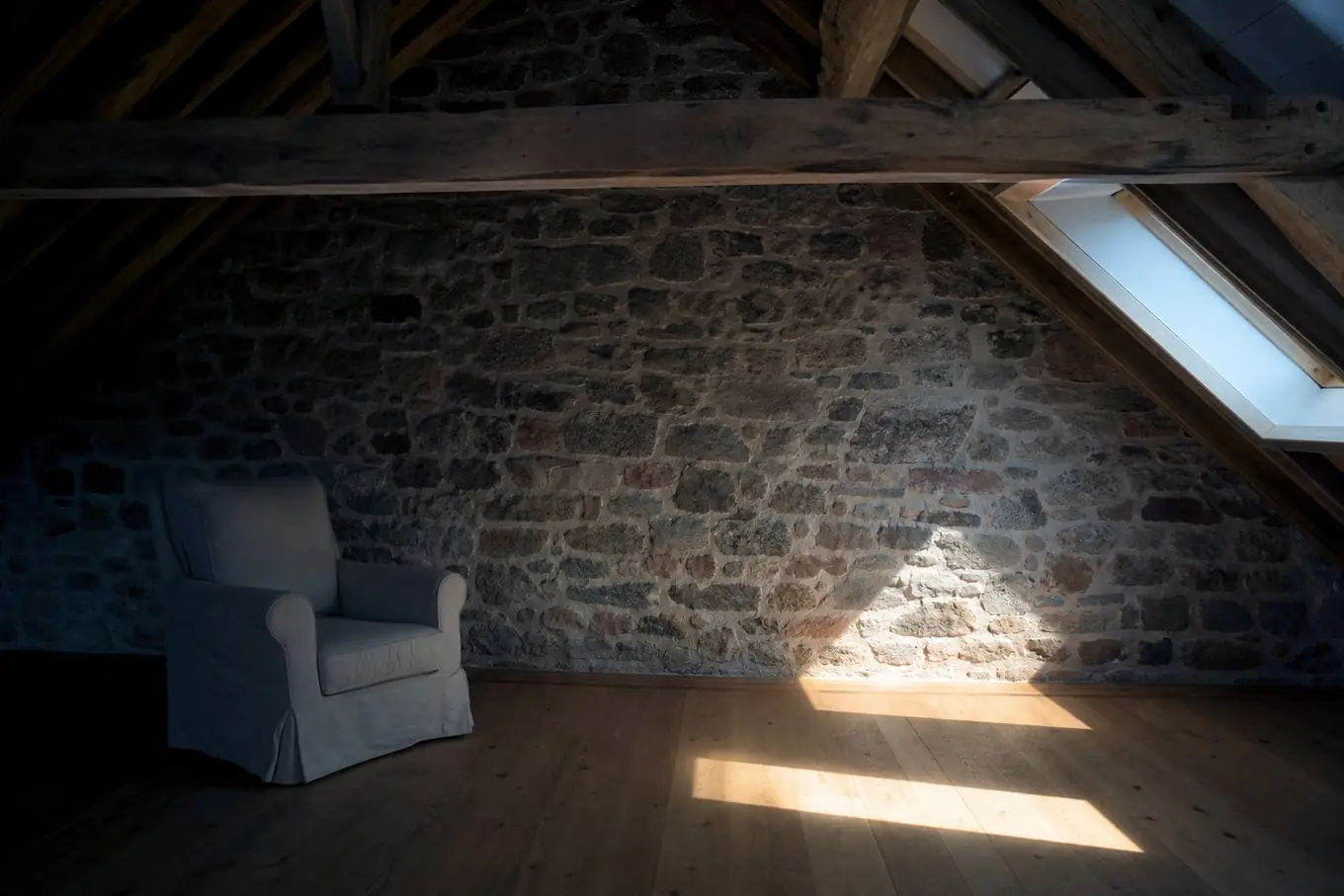Comble sous charpente de chêne, mur en moellons de granit et de grès rose, un rai de lumière entre par la fenêtre de toit et dessine un rectangle sur le plancher