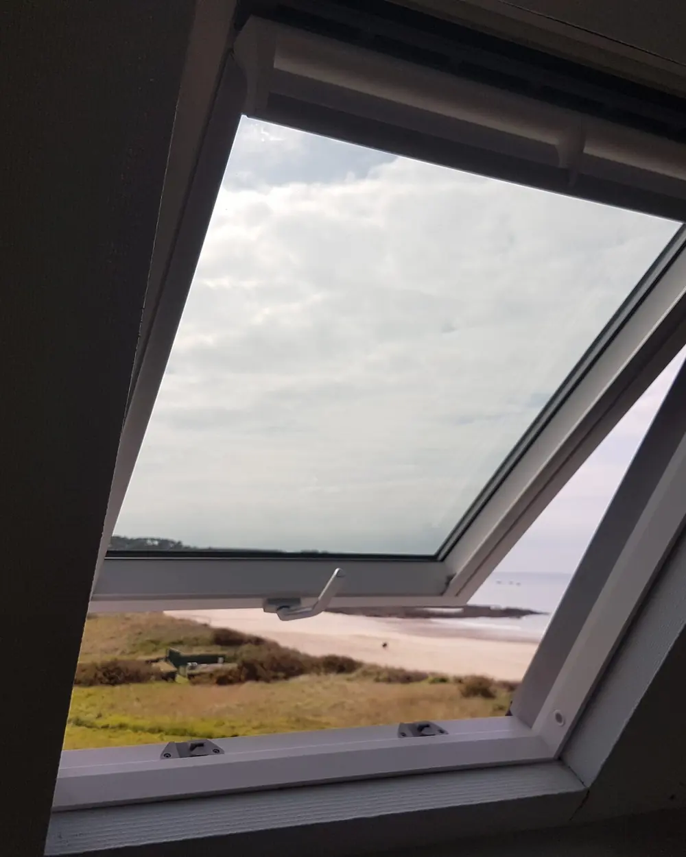 Fenêtre de toit blanche ouverte dans un rampant sombre : au-dehors, la lande, la plage et la mer
