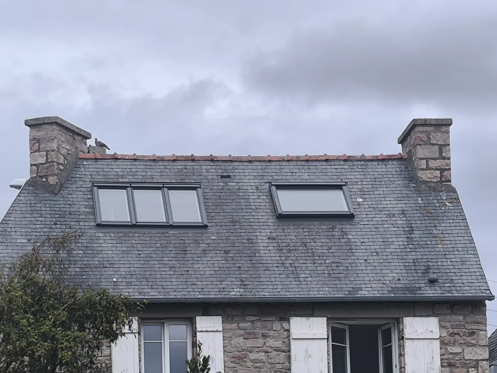 Pignon d'une maison bretonne en pierre, toiture en ardoise portant trois fenêtres de toit alignées d'un côté du pan et une quatrième de l'autre, deux souches de granit, ciel gris