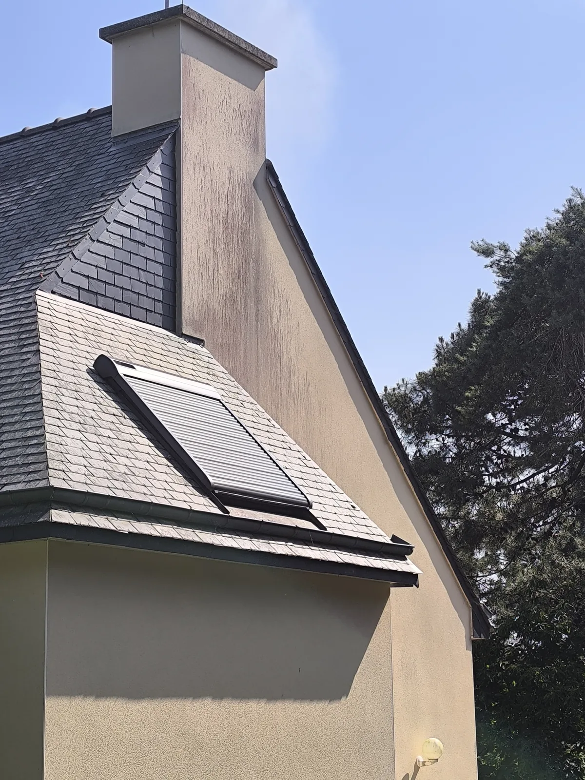 Volet roulant baissé sur une fenêtre de toit, dans la couverture en ardoise d'une lucarne, contre une souche de cheminée
