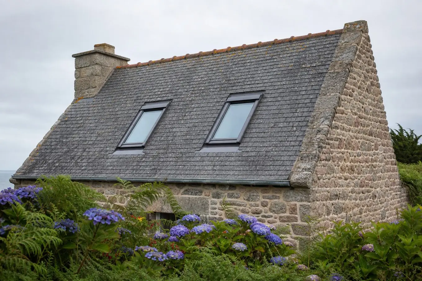 Maison de bord de mer en moellons de granit et de grès rose, toiture en ardoise portant deux fenêtres de toit, hortensias au premier plan