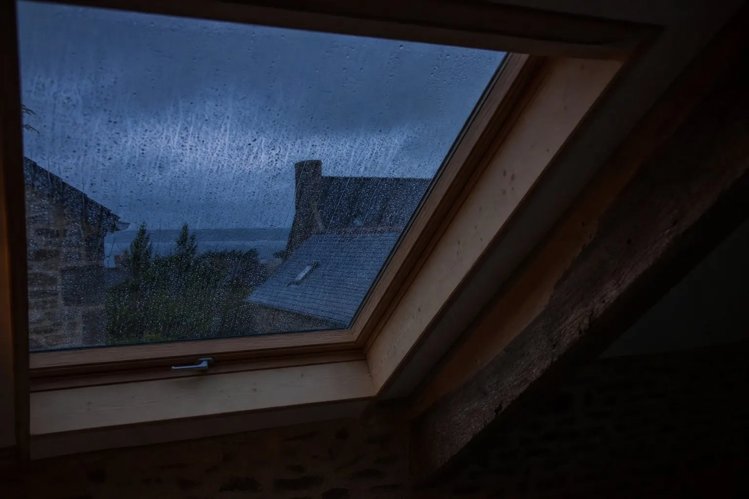 Fenêtre de toit vue de l'intérieur pendant une averse, ruissellement sur le vitrage, lumière bleue du dehors